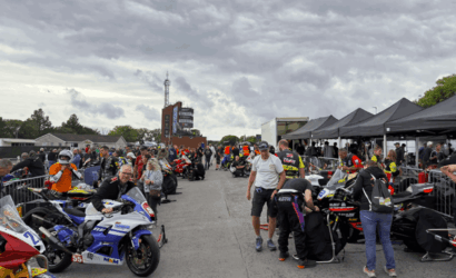 Crowd in a motorcycle paddock with racers and crews beside lined sport bikes under tents at a track event.