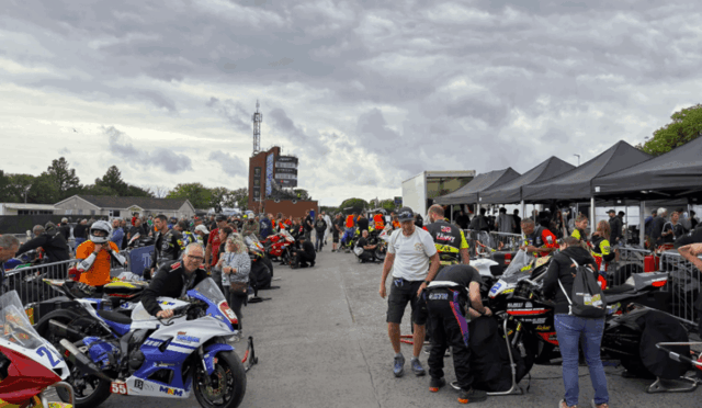 Crowd in a motorcycle paddock with racers and crews beside lined sport bikes under tents at a track event.