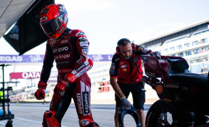 MotoGP rider in a red Lenovo Ducati suit walks from the pit lane as a crew member holds a helmet nearby next to a racing bike with sponsor logos.