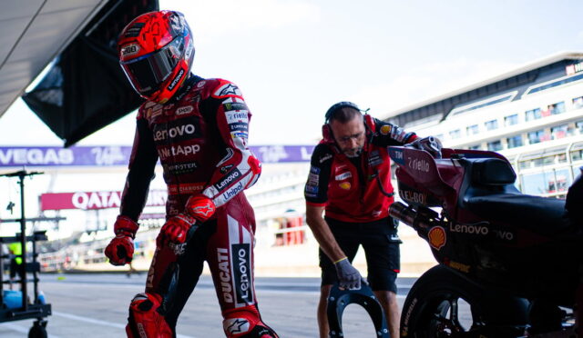 MotoGP rider in a red Lenovo Ducati suit walks from the pit lane as a crew member holds a helmet nearby next to a racing bike with sponsor logos.