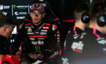 MotoGP rider in an Aprilia racing suit sits in the garage, talking with the crew; Monster Energy logos are visible in the background.