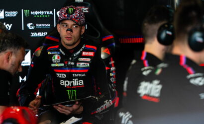 MotoGP rider in an Aprilia racing suit sits in the garage, talking with the crew; Monster Energy logos are visible in the background.