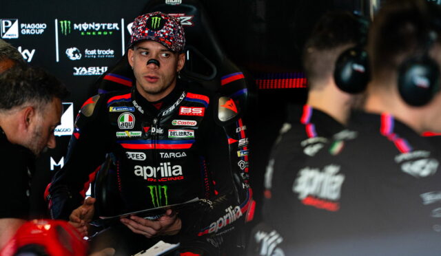 Motorsport rider in an Aprilia racing suit sits in the pit, talking with crew, Monster Energy logos in the background.