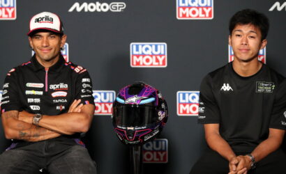 Two MotoGP riders in black team shirts sit with arms folded, a purple and black helmet on a stand between them, in front of a Liqui Moly backdrop.