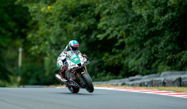 Motorcycle racer in full gear leaning into a curve on a race track with green trees in the background, helmet blue.