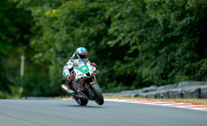 Motorcycle racer wearing a blue helmet leans into a turn on the track, number 30 on the bike, with green trees in the background.