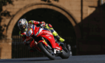 MotoGP rider in a red Honda bike leaning into a left-hand turn on a race track with a stone archway in the background.