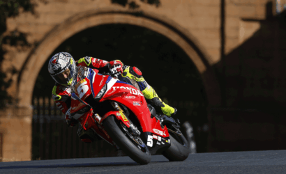 MotoGP rider in a red Honda bike leaning into a left-hand turn on a race track with a stone archway in the background.
