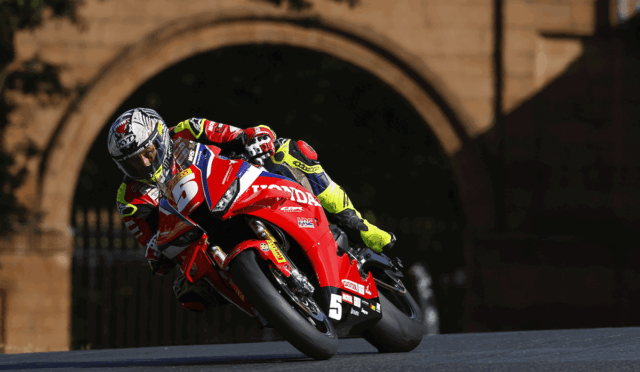 MotoGP rider in a red Honda bike leaning into a left-hand turn on a race track with a stone archway in the background.