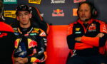 MotoGP rider in a Red Bull KTM suit sits in a racing chair beside a team member wearing a headset in the pit area with sponsor banners behind them.