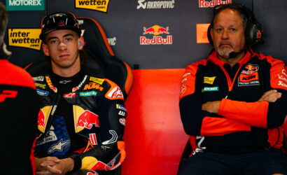 MotoGP rider in a Red Bull KTM suit sits in a racing chair beside a team member wearing a headset in the pit area with sponsor banners behind them.