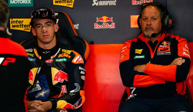 MotoGP rider in a Red Bull KTM suit sits in a racing chair beside a team member wearing a headset in the pit area with sponsor banners behind them.