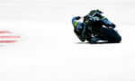 Motorcycle racer in a blue-green suit leaning into a sharp turn on a race track with red-and-white curb nearby.
