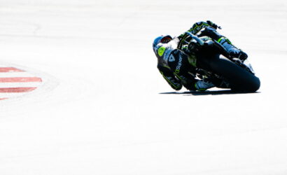 Motorcycle racer in a blue-green suit leaning into a sharp turn on a race track with red-and-white curb nearby.