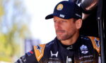 Male race car driver in a black and orange racing suit and cap, standing beside a vehicle, looking off to the side with a focused expression.