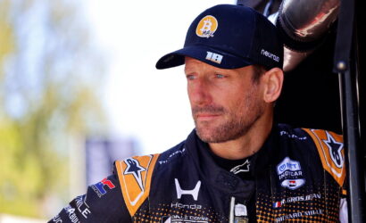 Male race car driver in a black and orange racing suit and cap, standing beside a vehicle, looking off to the side with a focused expression.