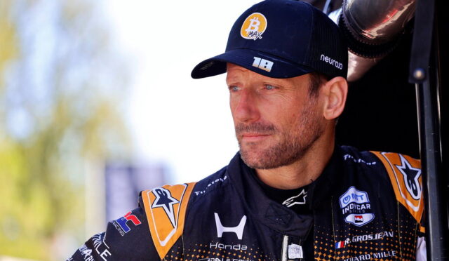 Male race car driver in a black and orange racing suit and cap, standing beside a vehicle, looking off to the side with a focused expression.