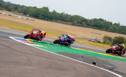 Three motorcycle racers lean into a curve on a race track, riders in red, blue, and dark gear close together.