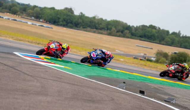 Three motorcycle racers lean into a curve on a race track, riders in red, blue, and dark gear close together.