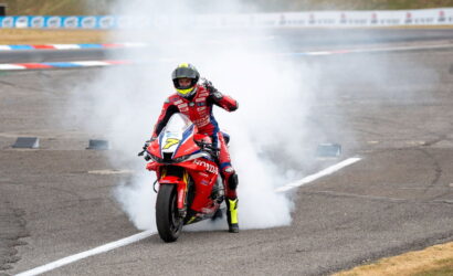 Motorcycle racer in red gear doing a burnout on a track, creating white smoke around a red Honda bike.