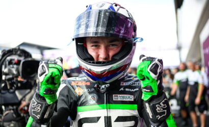 Motorcycle racer in colorful helmet and green gloves giving two thumbs up to the camera.
