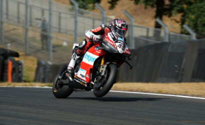 Motorsport rider leaning into a left turn on a red‑white racing bike with sponsor livery on track barriers in the background