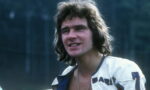 Young man with shoulder-length brown hair wearing a white leather jacket outdoors, smiling slightly.