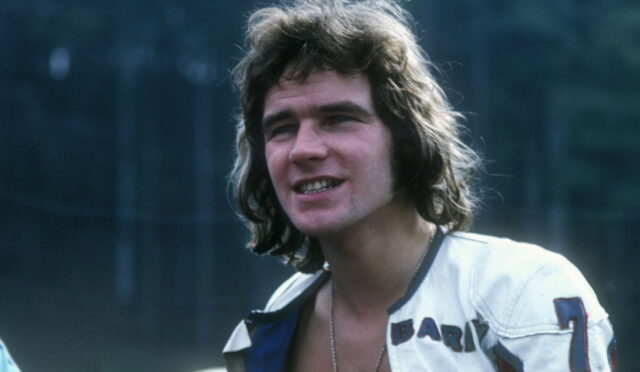 Young man with shoulder-length brown hair wearing a white leather jacket outdoors, smiling slightly.