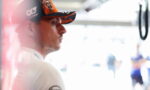 Side profile of a man in a black and orange AlphaTauri cap, looking focused in a bright pit area.