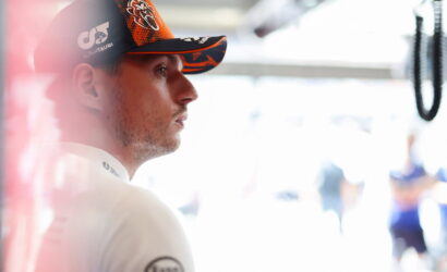 Side profile of a man in a black and orange AlphaTauri cap, looking focused in a bright pit area.