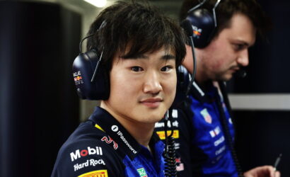 Young man in a dark blue racing jersey with sponsor logos, wearing a headset with microphone in a pit area.