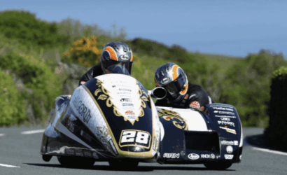 Sidecar motorcycle race in a sharp turn with two riders leaning into the curve; number 28 on the front and gold-black livery against a green, sunny backdrop.