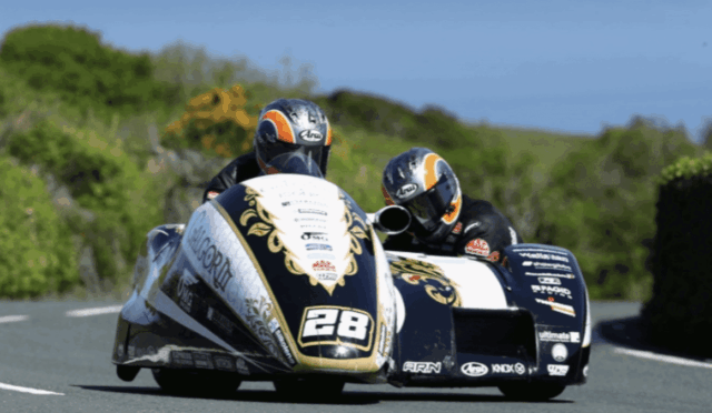 Sidecar motorcycle race in a sharp turn with two riders leaning into the curve; number 28 on the front and gold-black livery against a green, sunny backdrop.