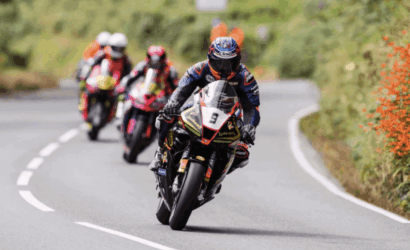 Motorcycle racer in blue gear leaning into a curve with a numbered bike (9) leading a group on a winding road outskirts.