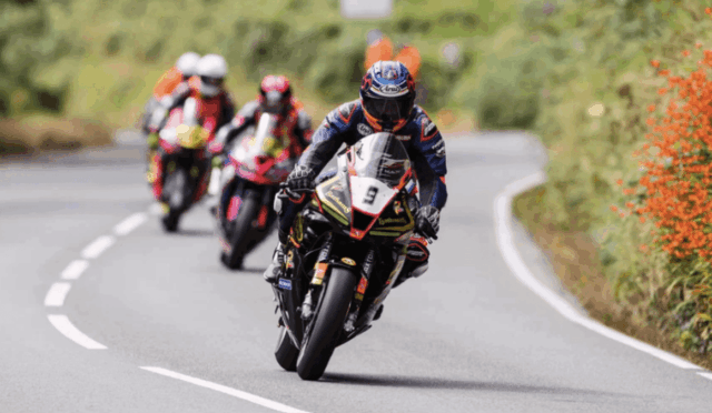 Motorcycle racer in blue gear leaning into a curve with a numbered bike (9) leading a group on a winding road outskirts.