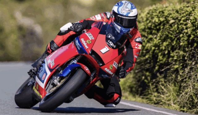 Racer on a red and blue motorcycle leaning through a turn on a paved road, helmeted and in full gear, hedge beside the track