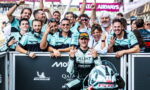 Racing team celebrates a podium moment with the rider in front, fans and teammates cheering behind a barrier at the trackactions.