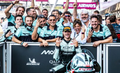 Racing team celebrates a podium moment with the rider in front, fans and teammates cheering behind a barrier at the trackactions.