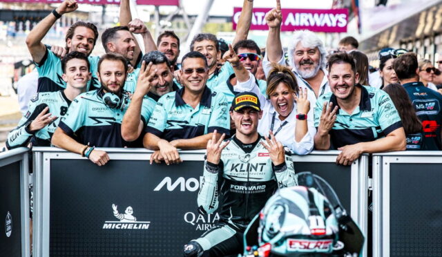 Racing team celebrates a podium moment with the rider in front, fans and teammates cheering behind a barrier at the trackactions.