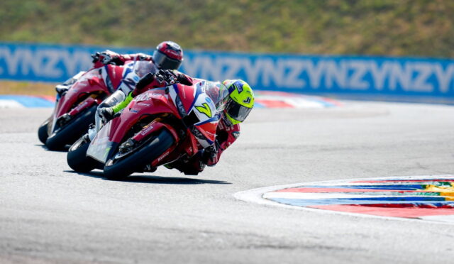 Three motorcyclists in red racing suits lean into a curve on a race track during a competition with blurred blue barriers in the background.