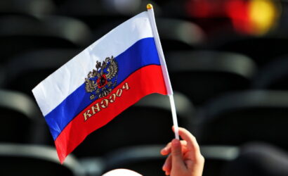 Hand holding a small Russian flag with the coat of arms against a dark blurred background.