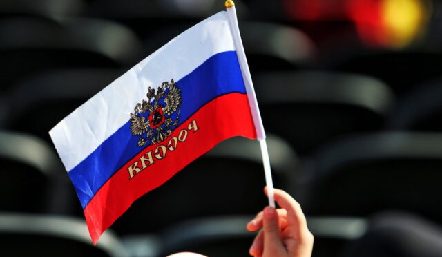 Hand holding a small Russian flag with the coat of arms against a dark blurred background.