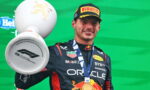 Race car driver on podium holding a large trophy with a gold medal around neck; wearing Red Bull suit and Pirelli cap, green backdrop visible.