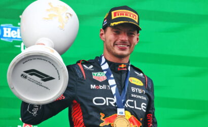 Race car driver on podium holding a large trophy with a gold medal around neck; wearing Red Bull suit and Pirelli cap, green backdrop visible.
