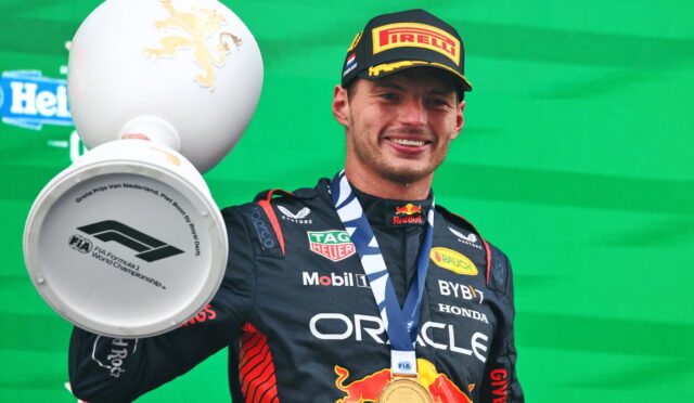 Race car driver on podium holding a large trophy with a gold medal around neck; wearing Red Bull suit and Pirelli cap, green backdrop visible.