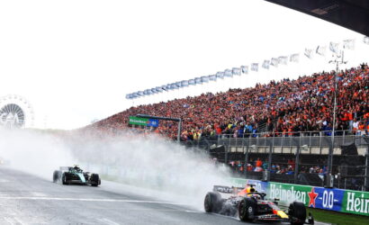 Two Formula 1 cars race past a packed grandstand as smoke trails behind them; Heineken banners line the track.