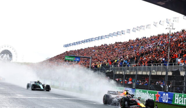 Two Formula 1 cars race past a packed grandstand as smoke trails behind them; Heineken banners line the track.