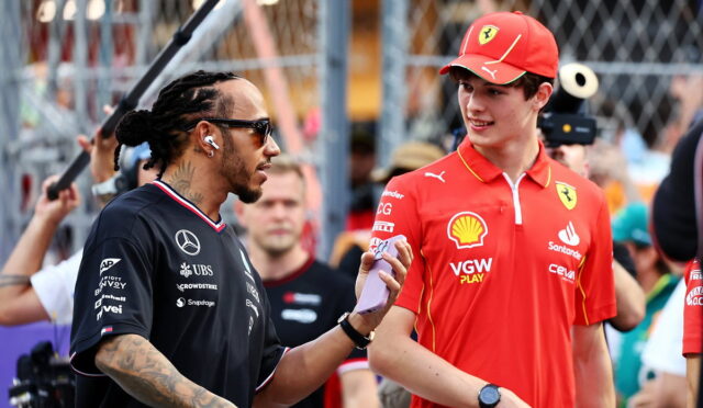 Man with braided hair in a black polo talks to a young Ferrari team member in a red shirt and cap at a gated event behind a crowd of spectators.