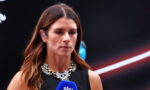 Female reporter with a blue Sky microphone, speaking at a press event, wearing a black sleeveless top and a jeweled necklace.