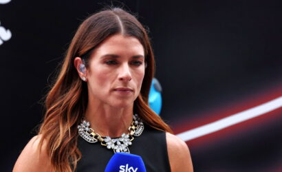 Female reporter with a blue Sky microphone, speaking at a press event, wearing a black sleeveless top and a jeweled necklace.